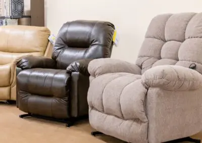 Row of three showroom recliners: beige leather recliner at left, dark brown leather recliner in center, and light gray fabric power recliner with padded tufted back and armrests at right.