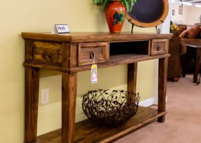 Rustic wooden console table with lower shelf and small drawers displayed against a pale yellow wall in a showroom, topped with a red ceramic vase with green plant and decorative platter, price tag hanging from a drawer and metal decorative bowl on the bottom shelf, staged on beige carpet