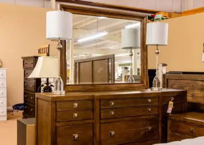 Rustic brown wooden dresser with nine drawers and square metal knobs, matching framed mirror on top reflecting showroom lights, two clear-glass table lamps with white shades flanking the mirror, and a price tag visible on the right drawer in a furniture store setting