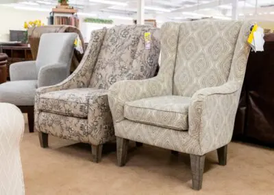 Two patterned upholstered accent chairs — one with beige geometric fabric and one with floral print — displayed side-by-side on a showroom floor with price tags attached and other furniture visible in the background