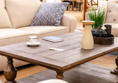 Brown wooden coffee table styled with a beige ceramic vase, a black metal tray holding decorative items and faux grass, a teacup on a saucer, and a closed blue notebook between two tufted beige sofas in a furniture showroom setting