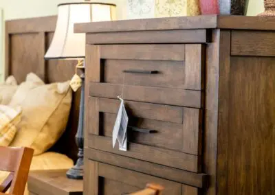 Brown wooden three-drawer nightstand with modern black handles and a price tag hanging from the middle drawer, shown in a furniture showroom next to a lamp, beige throw pillows and a wooden chair.
