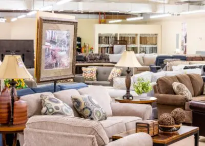 Beige corduroy loveseat with patterned throw pillows and wooden coffee table topped with decorative wicker balls and small box, surrounded by floor lamps, framed artwork, and multiple sofas in a bright furniture showroom