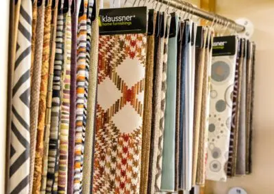 Fabric sample swatches hanging on a metal display rod in a home furnishings showroom, showing patterned and solid upholstery textiles including a prominent brown-and-orange geometric klaussner sample.