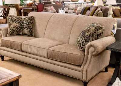 Beige three-seat rolled-arm sofa with patterned throw pillows displayed in a furniture showroom on a carpeted floor, surrounded by side tables and lamps