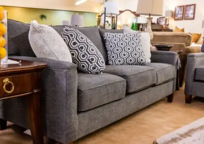 Gray three-seat upholstered sofa in a showroom setting with patterned and textured throw pillows, a wooden side table with a glass vase of oranges, matching armchair and other furniture displayed on beige carpet