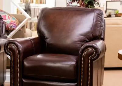 Brown leather swivel armchair with rolled arms and brass nailhead trim displayed on showroom carpet with sofas, fabric samples and plants in the background