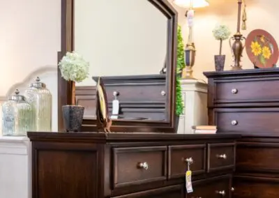 Dark wood bedroom dresser with arched mirror, silver knobs, bedside lamp and decorative vases on top, styled in a furniture showroom setting.