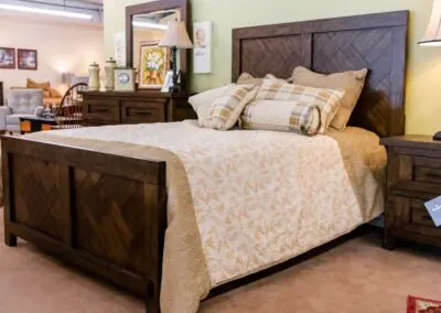 King-sized rustic dark-wood bed with patterned beige bedspread, plaid and bolster pillows, matching nightstand and dresser in a furniture showroom setting