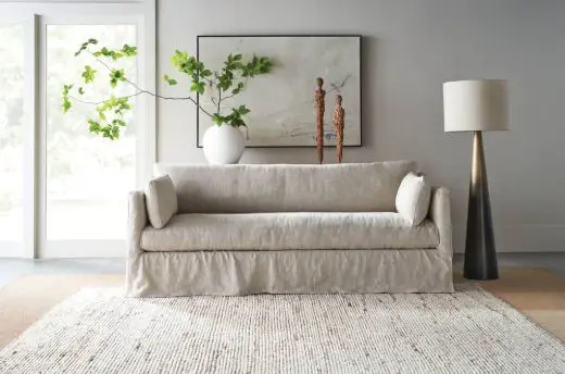 Neutral beige slipcovered sofa centered on a textured rug in a bright living room with a tall floor lamp to the right, a white vase with leafy branches and sculptural wood figures on a simple console behind the sofa, and a large framed minimalist artwork on a light gray wall.
